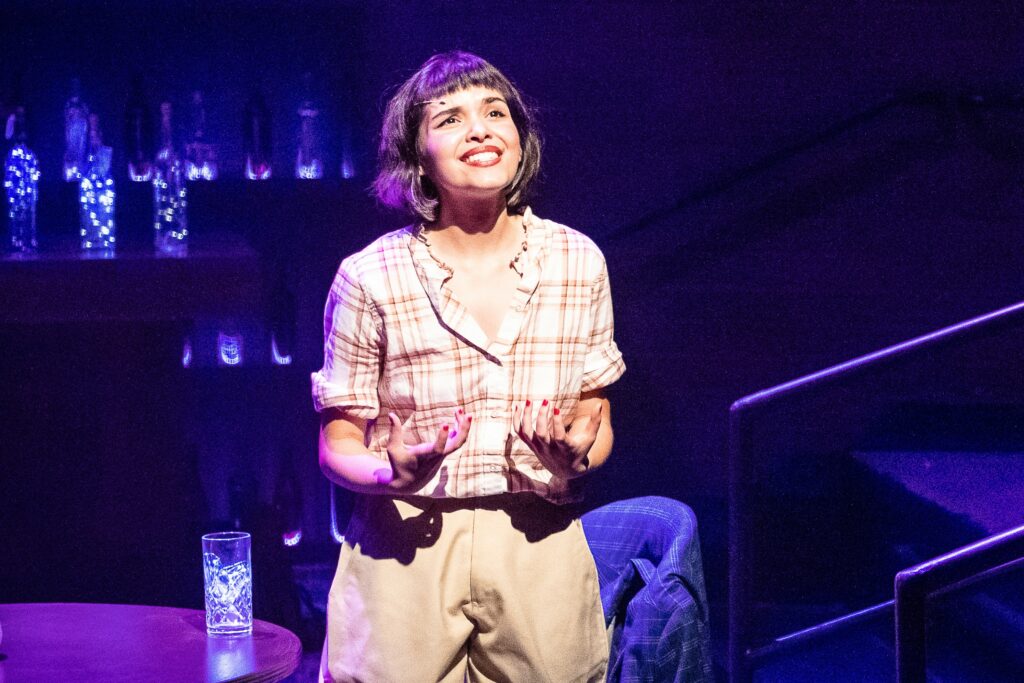 Woman on stage looking expectantly with purple background and glass on table