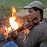 Photo of Miriam Oommen playing fiddle with campfire in background