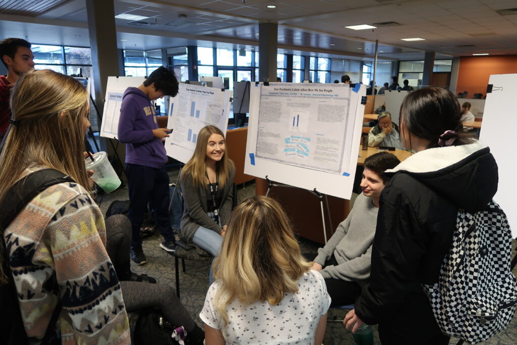 Group of people sitting and standing around a research poster listening to one person explain it 