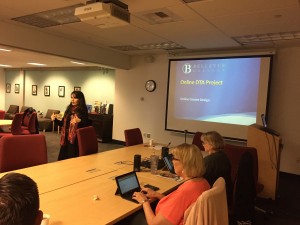 Ekaterina stands in front of the presentation screen in Faculty Commons and gestures with her hands. She is wearing a black dress, and is standing beside one of the red office chairs in Faculty Commons. On the presentation screen behind her is the title slide from the PowerPoint. The slide is Bellevue Blue, and says, "Online DTA Project."