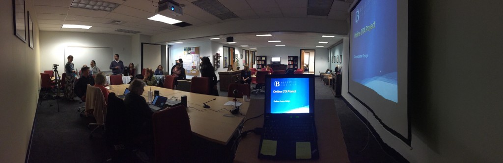 A panorama shot of Faculty Commons that encompasses the whole room and all the people attending. Sue Cox is in the left corner, standing and speaking to the room. In the right corner of the picture is the presentation screen with the words, Online DTA Project, as the title of the presentation.