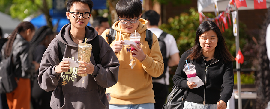 Three students in hoodie sweatshirts at an outdoor international Night Market event enjoying boba tea in the sunshine.