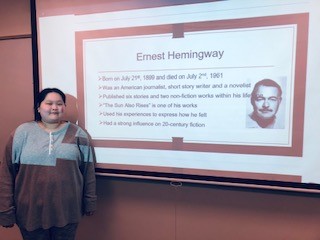 young adult female stands in front of a projecter screen with an image of ernest hemingway