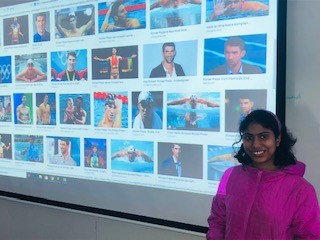 Young adult female stands in front of a projector screen with images of Michael Phelps