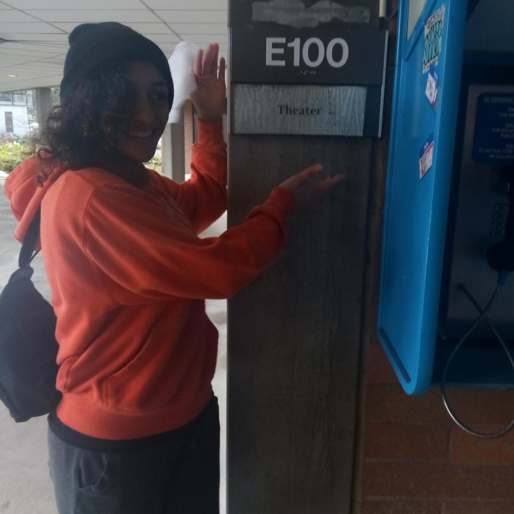 Female student poses in front ofa building, showing off the theater ign