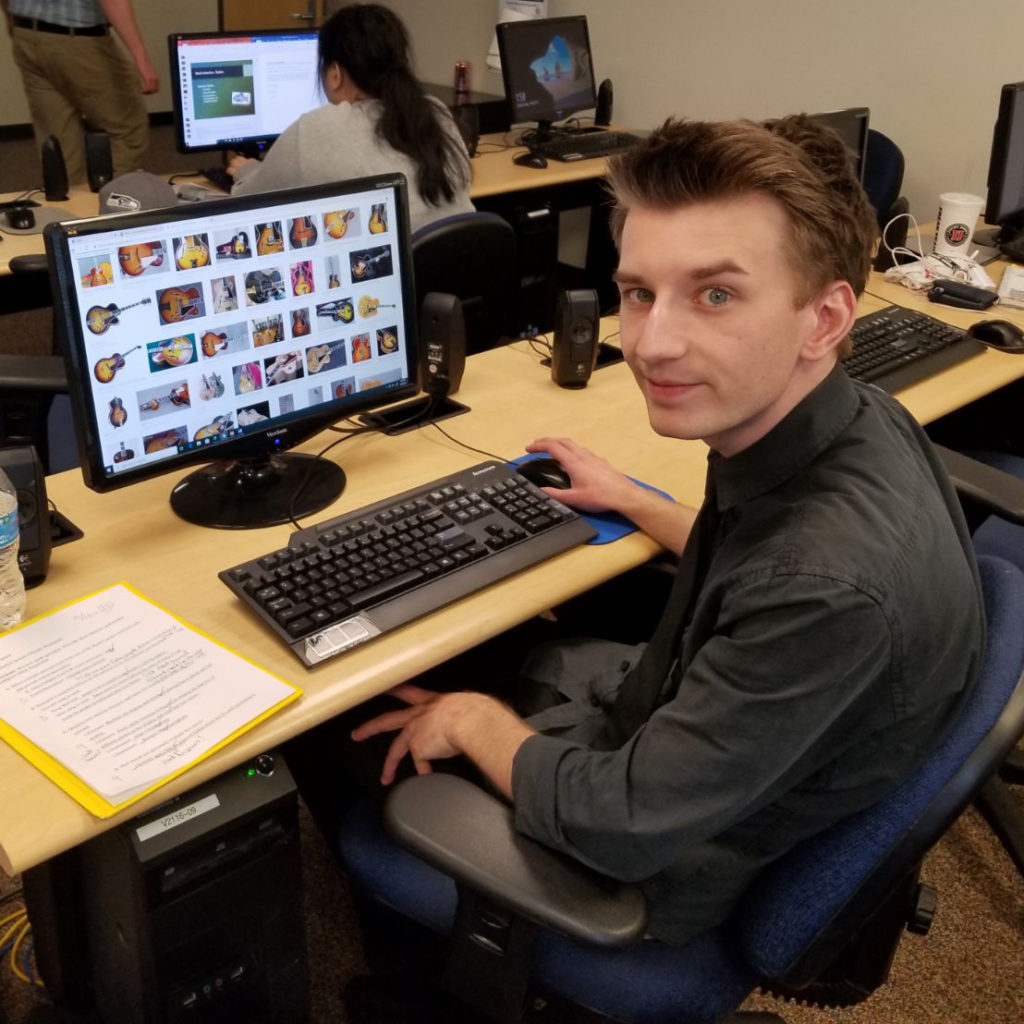 a male student sits at a computer looking at images of guitars