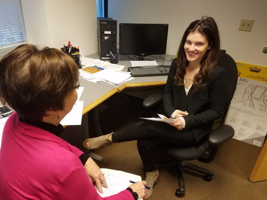 young adult woman sits dressed in business casual clothing sits at a desk being intrviewed by another adult woman. 