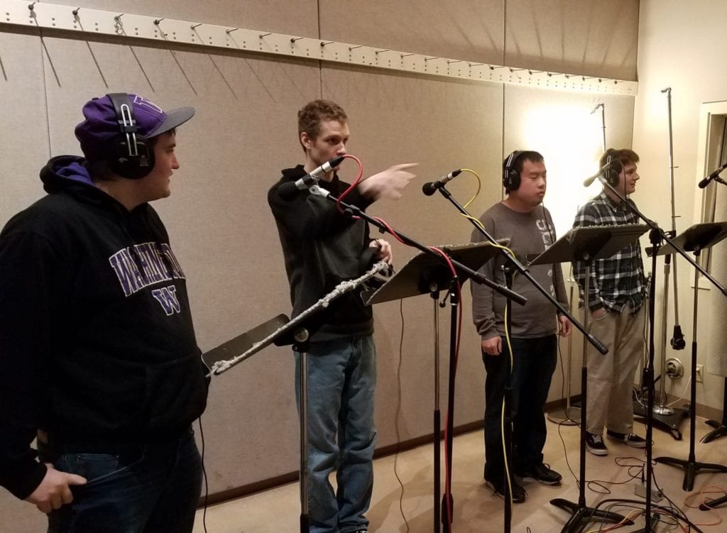 Four males stand at microphones in a studio