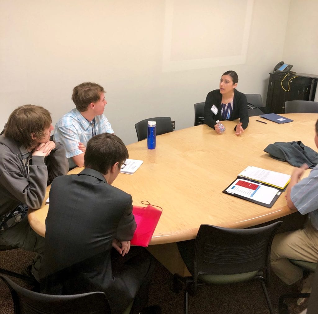 group of four young men sit at a table and listen to a young professional business woman at a conference table