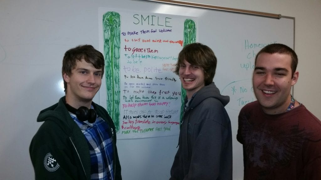 Three young adult male students stand in front of a poster that says "Smile"