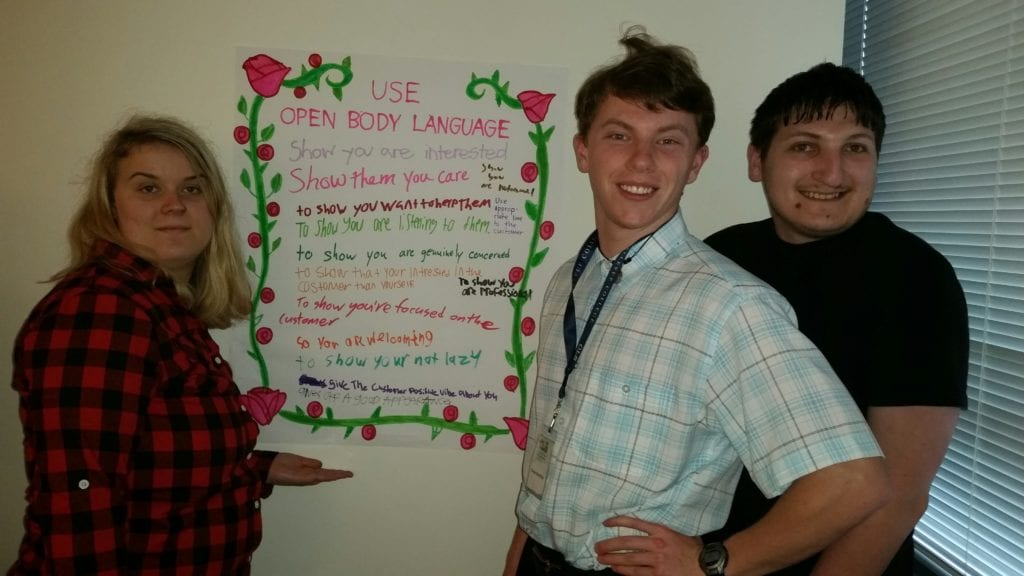 three young adult students smile in front of poster