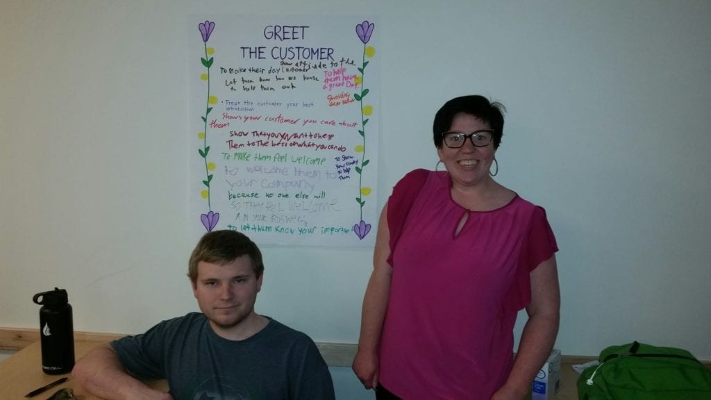 two young adult students stand in front of a poster explaining greeting customers