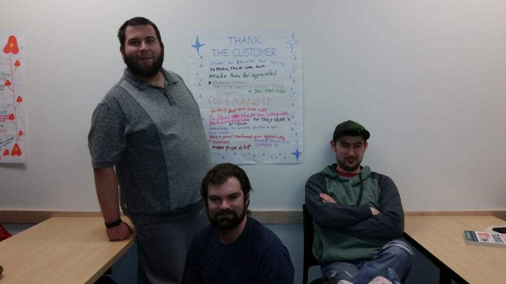 three young adult male students pose with a thank the cusomter poster