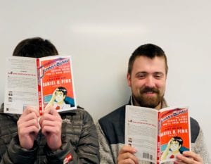 two young adult men reading a book, one using the book to cover his face, the other is smiling