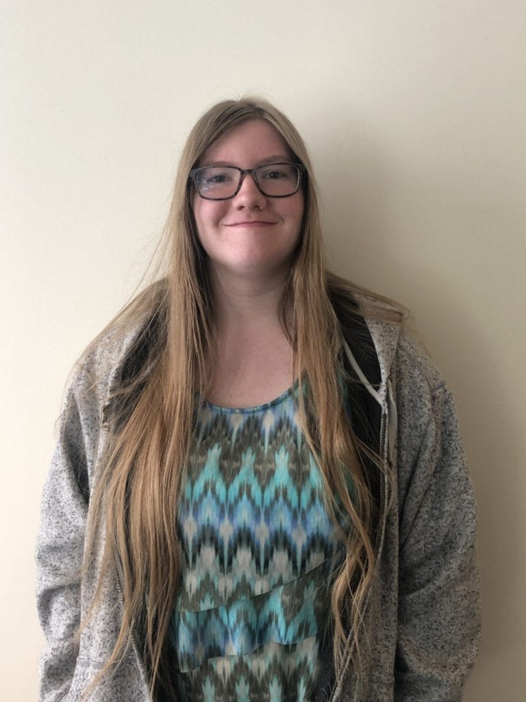 Young adult lady with long blong hair and glassesin grey sweater and brightly colored top smiles at the camera