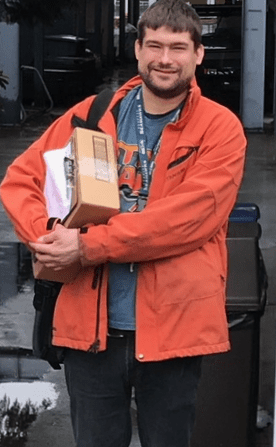 young man in orange coat stands in front of delivery area of building holding mail