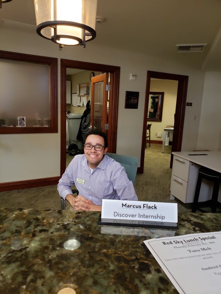 young man sits behind receptionist desk smiling