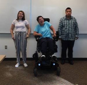 Three young adults students smile together in front of a whiteboard at the front of a college classroom. 