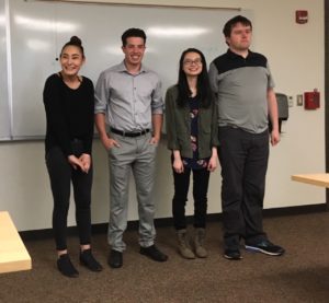 Four young adult students stand at the front of a college classroom smiling. 