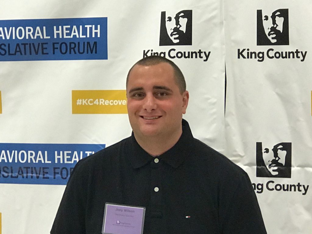 young man stands in front of King County Behavioral health forum backdrop