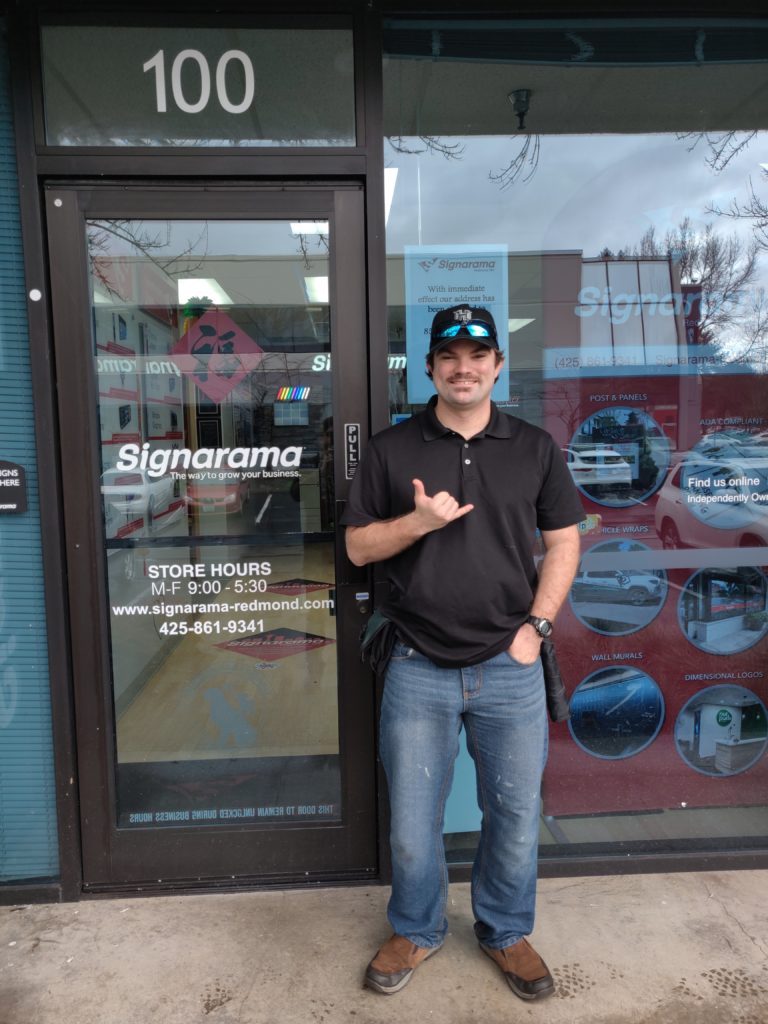 young man holds up "hang loose" hand signal in front of grpahic design shop