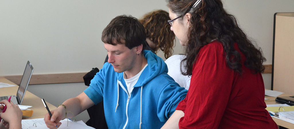 OLS Student works at a table and a faculty member stands near them helping them with their classwork.