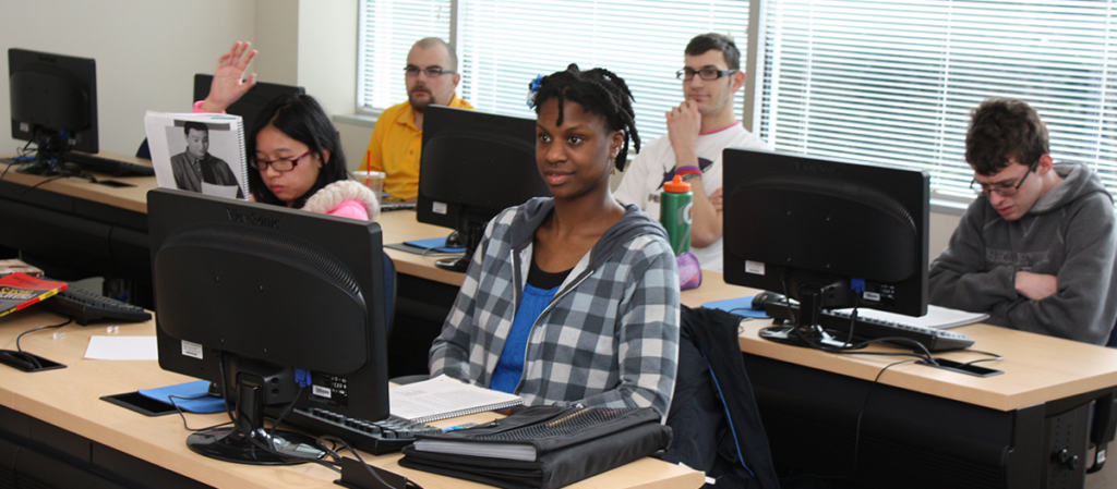 Five OLS Students sit at their desks in class listening to an instructor and one student has their hand raised to ask a question