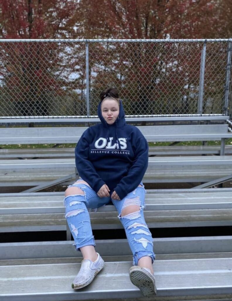 a young woman wearing an OLS Bellevue College sweatshirt sits on bleachers outside on a spring day