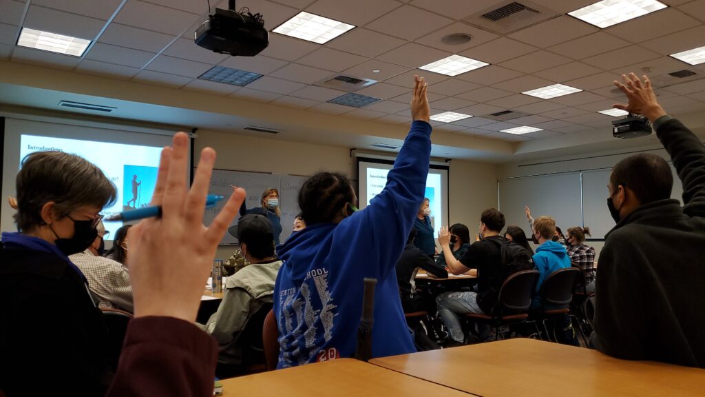 A group of college students in masks sit at desks in an indoor classroom, raising hands during a presentation. 