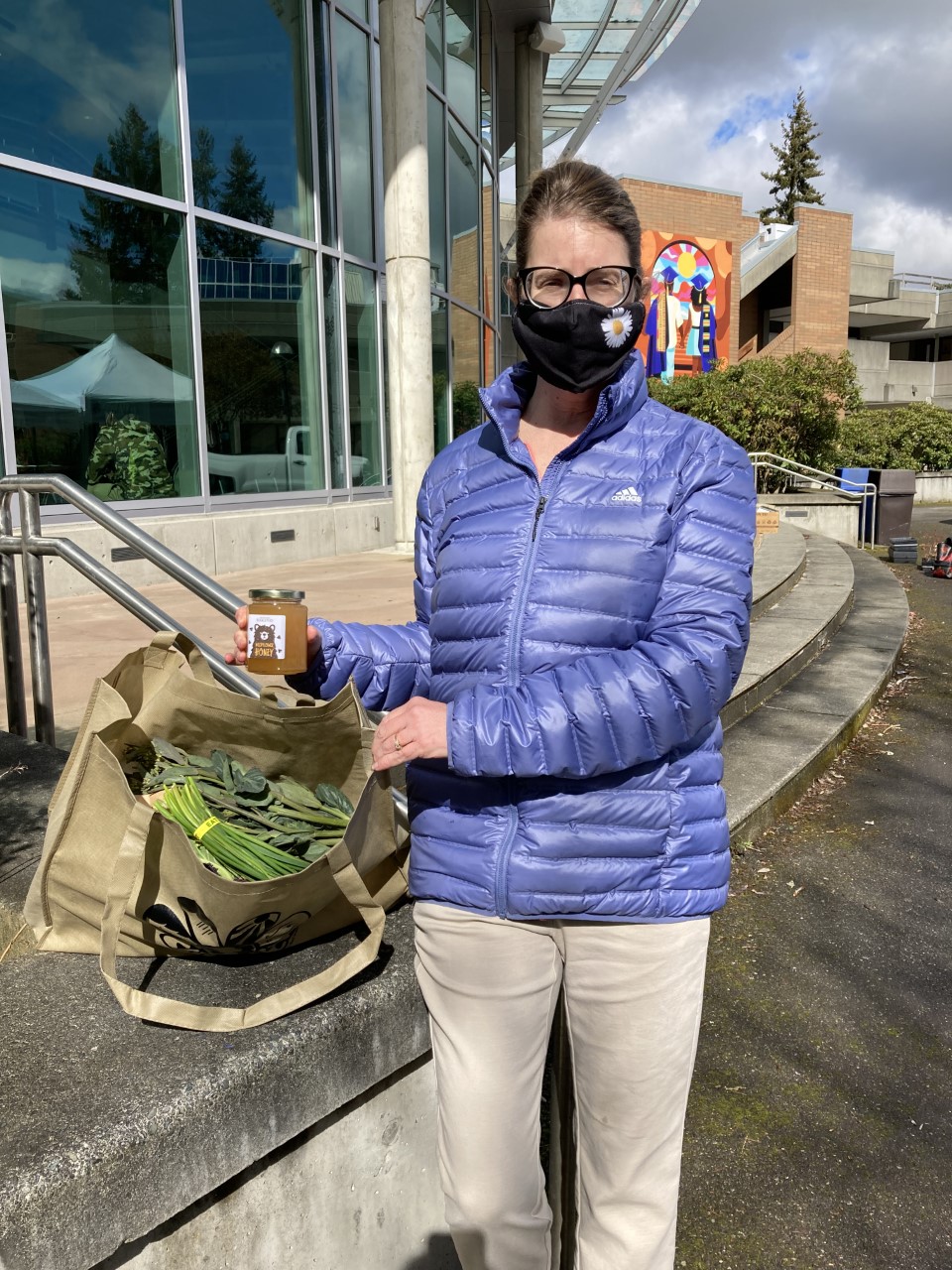 A woman in a mask outside on a college campus holds a jar of honey and a bag of produce.