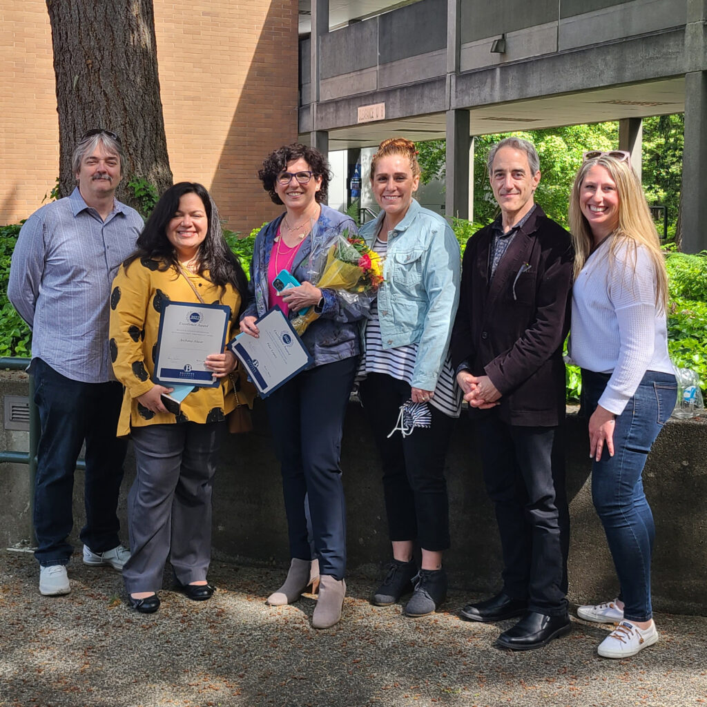 Six adults pose for a group photo outside on a college campus