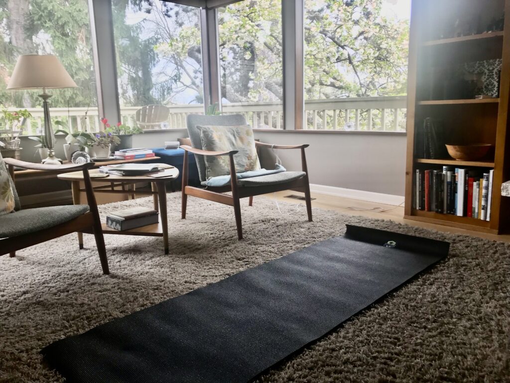 A yoga mat lays on the floor of a living room with large, sunny windows 