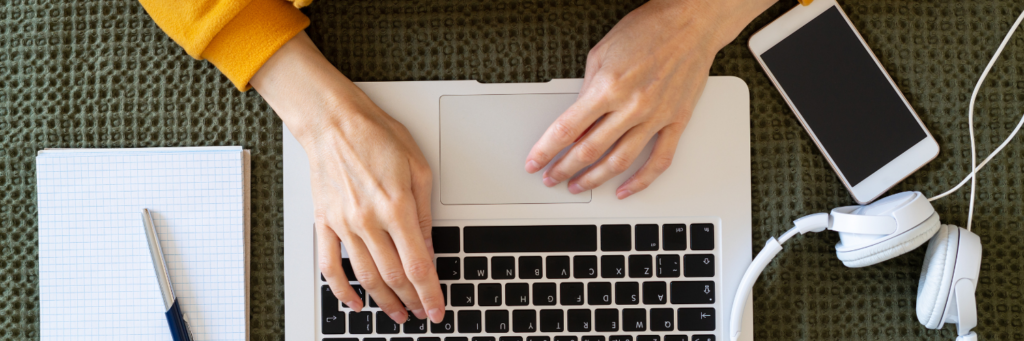 Image of person's hands working on a laptop with a mobile phone, headphones, pen and notebook nearby.