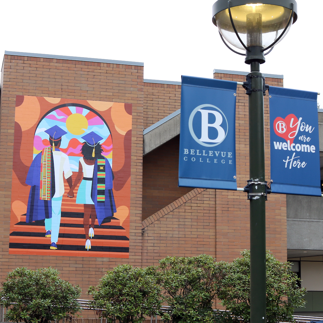 Unity Mural on the campus of Bellevue College, with a Bellevue College banner in the foreground that states, "You are welcome here". Mural depicts two Black students wearing graduation regalia as they walk up a staircase.