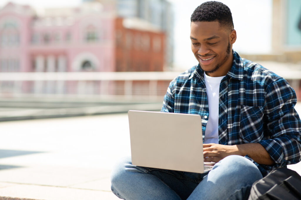 African American/Black student smiling and working on laptop outside on a sunny day.