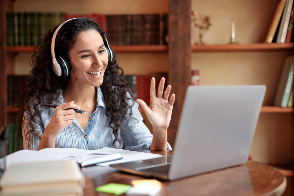 Smiling female student wearing headset is working on laptop and appears to be communicating with someone via video chat.