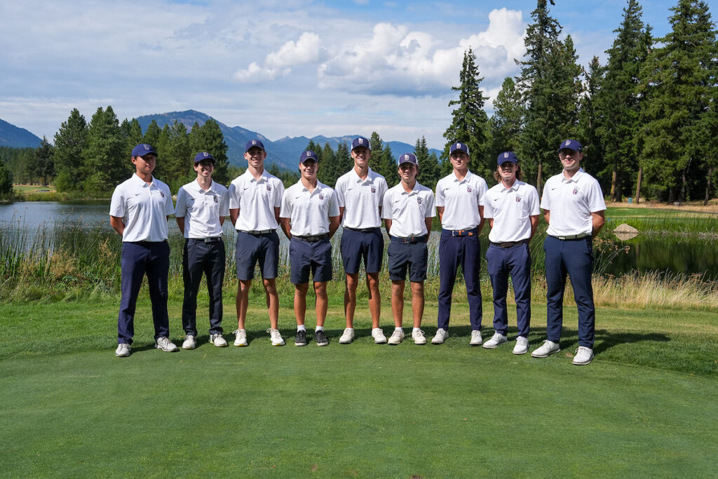 Men's golf team group, standing on the green in front of a large pond with mountains and clouds in the distance.