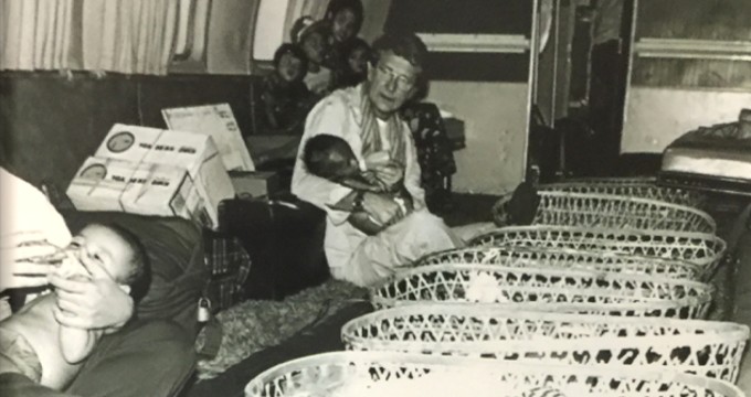Black and white photo of a male caretaker carrying a baby in his arms and multiple Cambodian orphans in the background