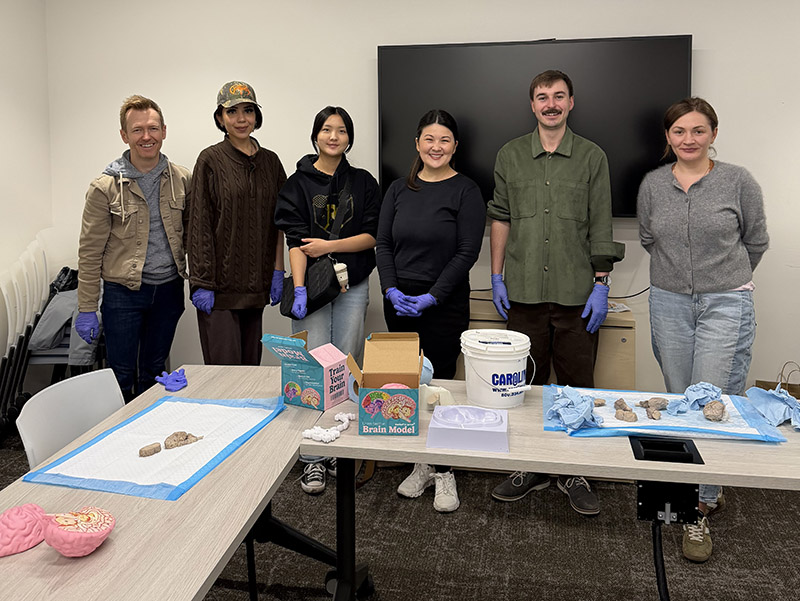 A group of people stands in a classroom behind tables displaying brain models and lab materials used for a hands‑on activity.