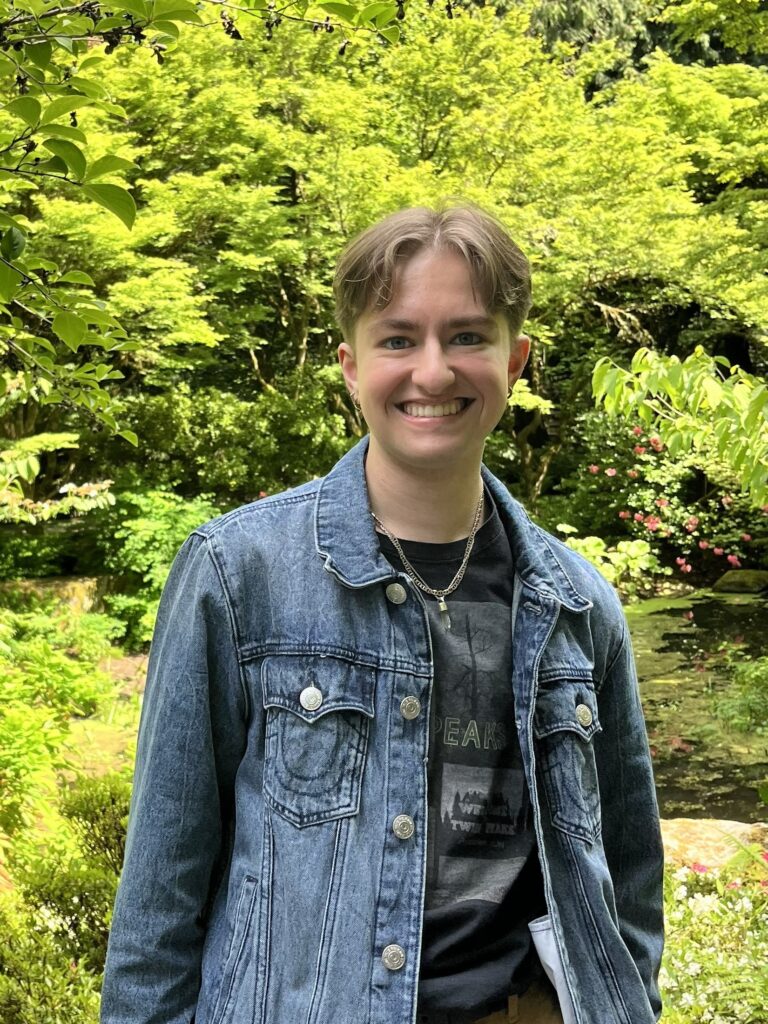 White nonbinary person smiling with short brown hair and blue eyes. They are wearing a Twin Peaks graphic tee shirt and a denim jacket with foliage in the background.