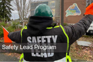 person with orange flags, jacket with white text reads Safety lead, white text on button reads Safety Lead Program