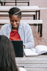 A student studies outside with a laptop, a notebook and another student.