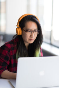 A student wearing headphones works on a laptop by a window.