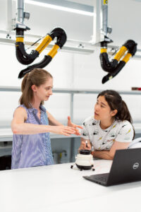 A professor and student discuss robotics in a lab, with a lapto and robot in front of them.