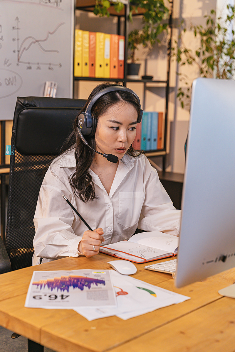 A woman in a headset works on a computer while consulting graphs.