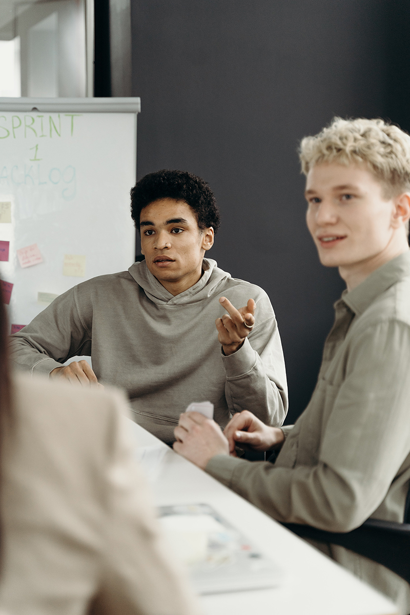 Two students, with a whiteboard and notes behind them, listen to another speaking.