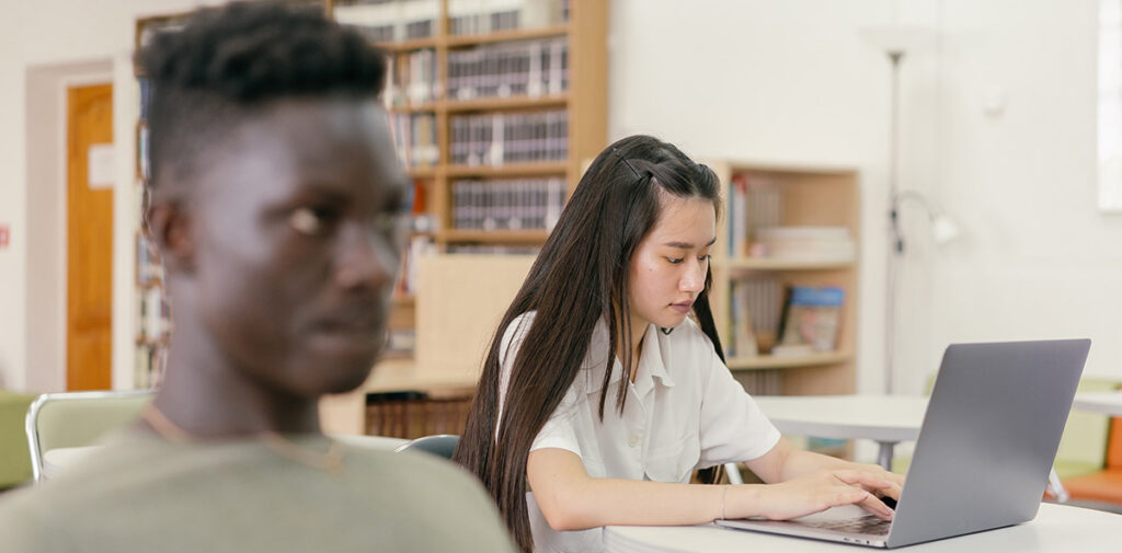 Two students, one with a laptop, listen and take notes in class.