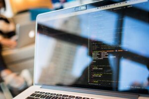 Open laptop with programming script on screen in foreground with blurred background image of office activity