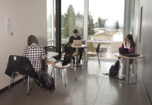 3 students studying at separate small tables. In the background there is a large window with a view of trees and surrounding rooftops.