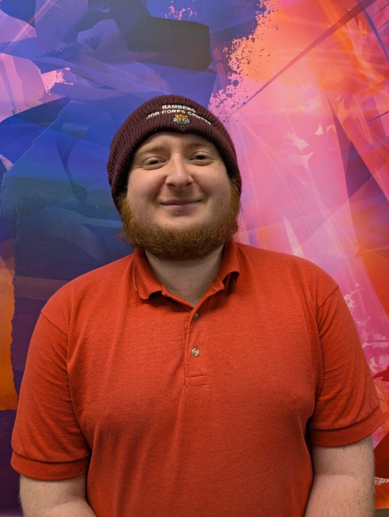 A person wearing a maroon beanie and a red polo shirt stands in front of a colorful abstract background with shades of purple, pink, orange, and blue. They are smiling softly at the camera.